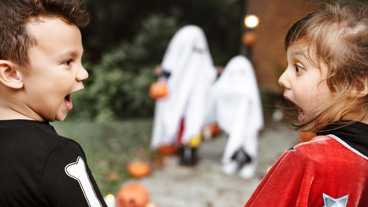 Two excited children (skeleton and red cape) with ghost-hooded kids and pumpkins in background outdoors.